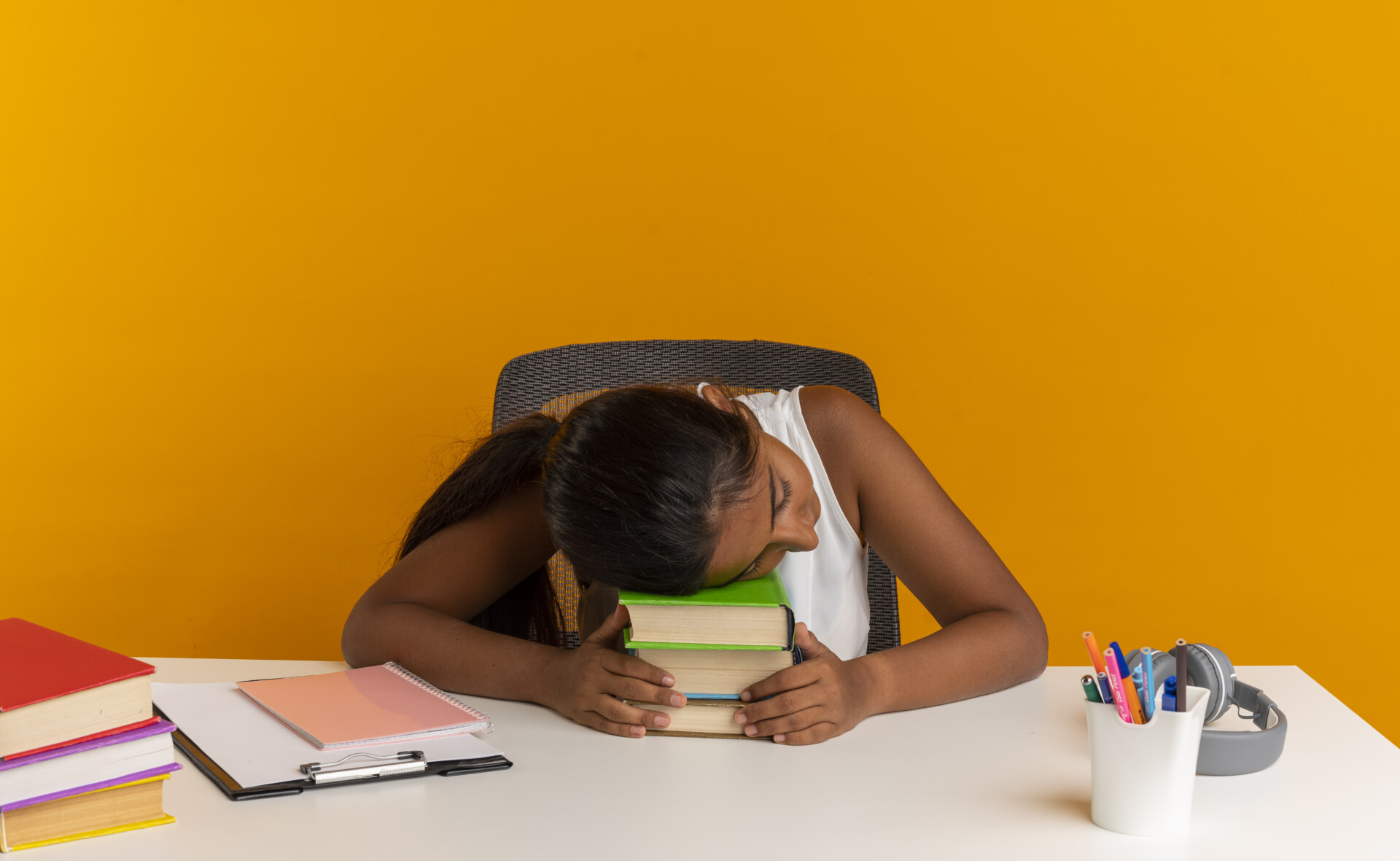 young schoolgirl sitting at desk with school tools putting head on books isolated on orange background