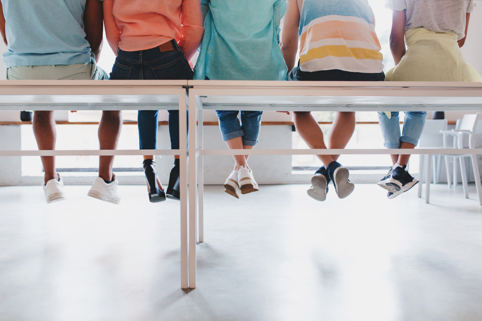 Indoor photo from back of student's leg in stylish colorful shoes. Portrait of young people sitting on white bench and resting after long walk outside.